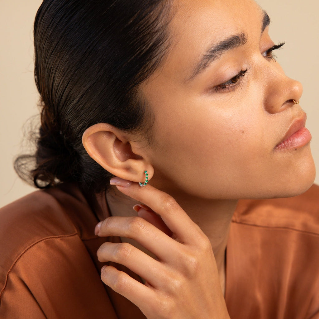 A woman with sleek hair wears elegant Inside Out Emerald Huggies, touching her ear as she gazes thoughtfully to the side.