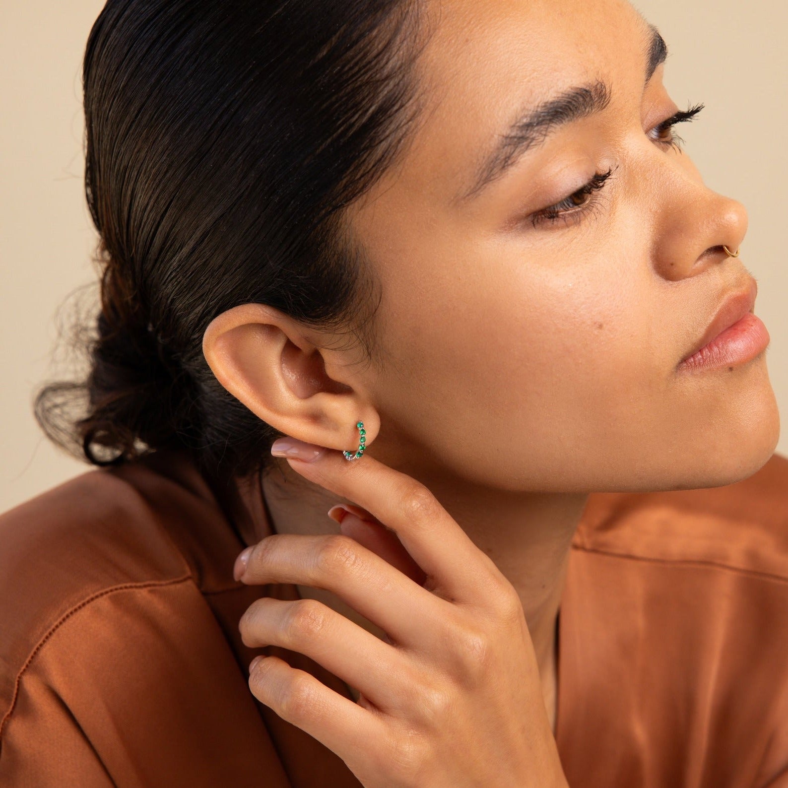 A woman with sleek hair wears elegant Inside Out Emerald Huggies, touching her ear as she gazes thoughtfully to the side.