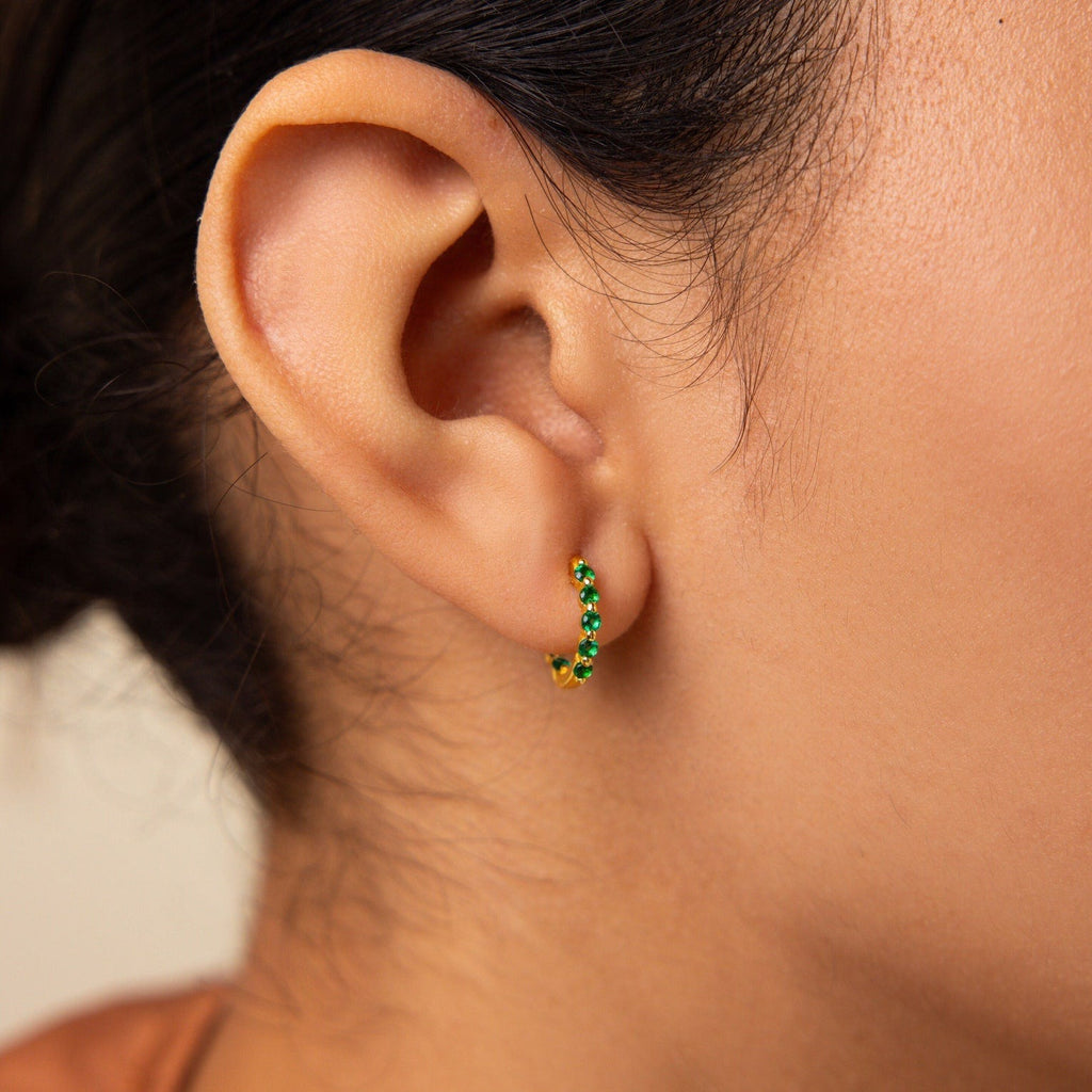 Close-up of a woman's ear wearing the Inside Out Emerald Huggies, small gold hoop earrings with green gemstones, showcasing elegant birthstone jewelry.