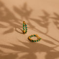 The Inside Out Emerald Huggies—gold hoop earrings with green gemstones—rest on a beige surface with leafy shadows, offering a stylish way to add elegant birthstone jewelry to your look.