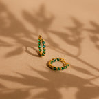 The Inside Out Emerald Huggies—gold hoop earrings with green gemstones—rest on a beige surface with leafy shadows, offering a stylish way to add elegant birthstone jewelry to your look.