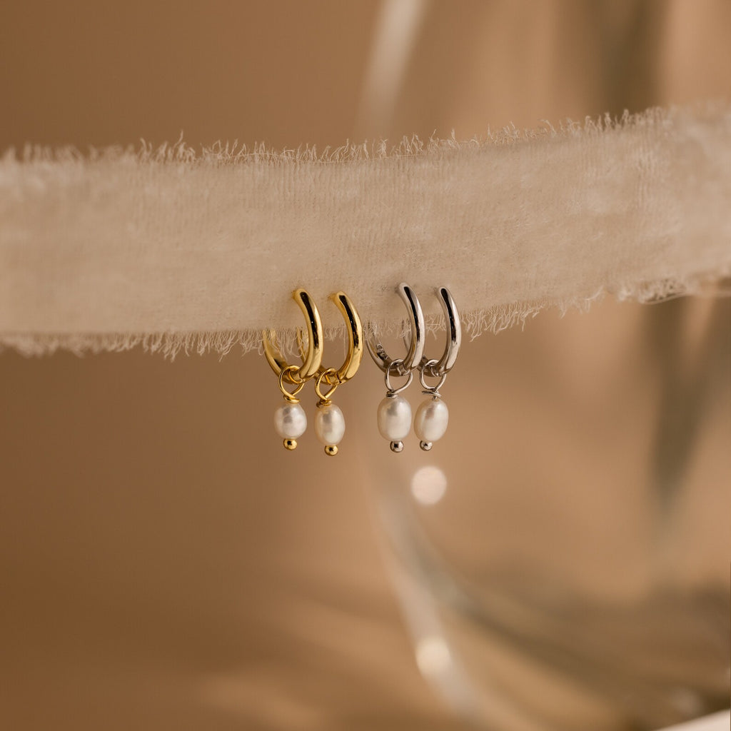 Close-up of Gold and Silver Dainty Pearl Huggies hanging on a beige fabric. 