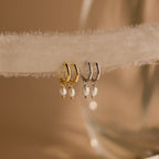 Close-up of Gold and Silver Dainty Pearl Huggies hanging on a beige fabric. 