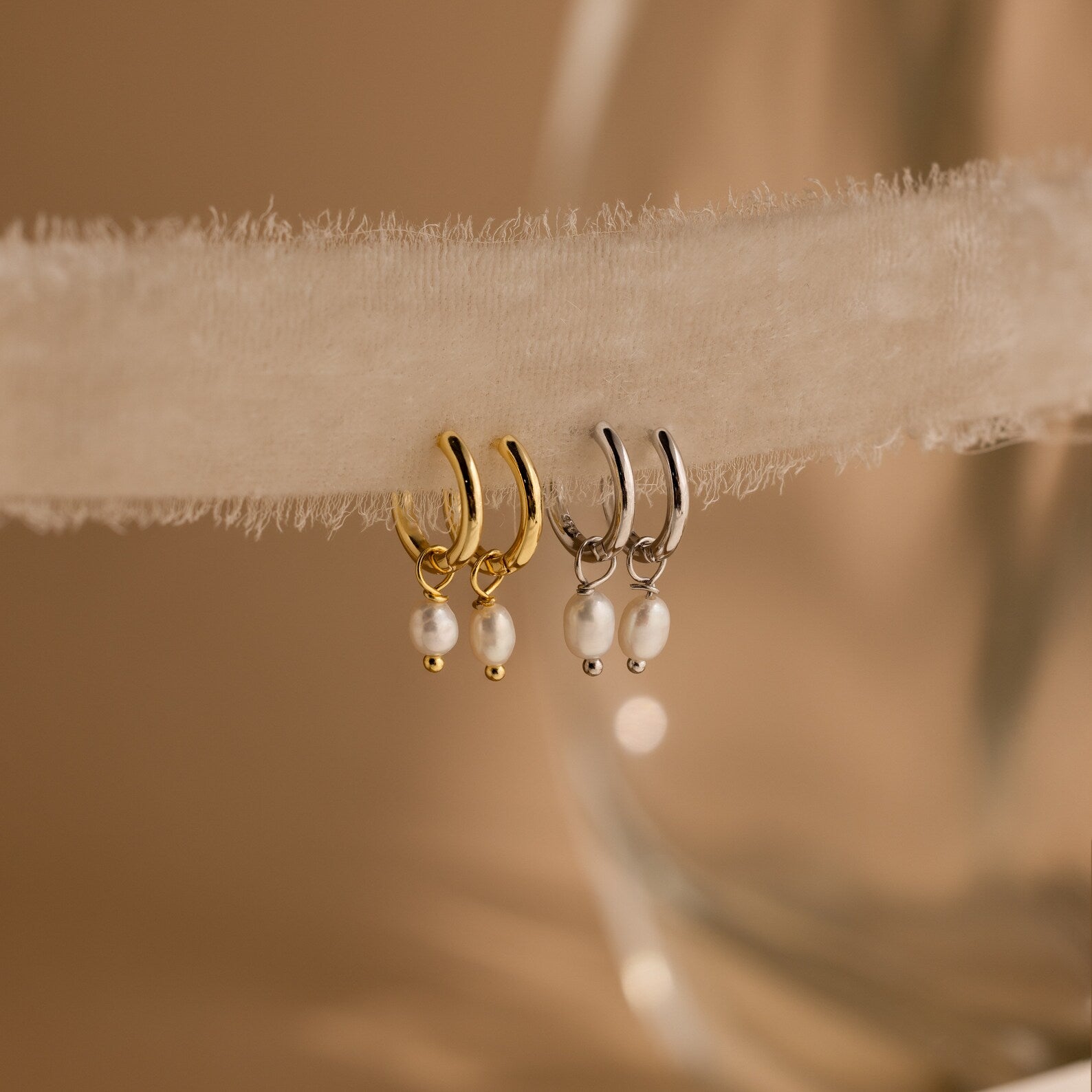 Close-up of Gold and Silver Dainty Pearl Huggies hanging on a beige fabric. 
