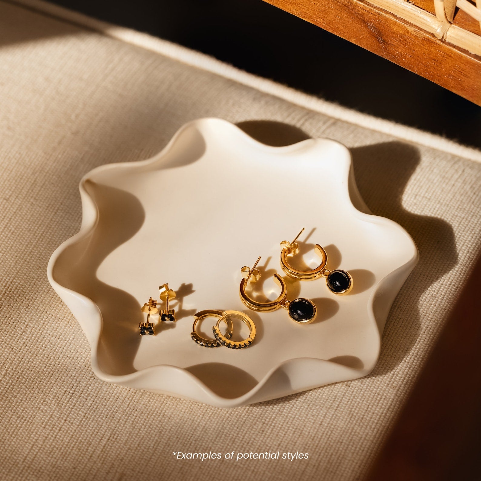 A wavy-edged ceramic dish holds the elegant Mystery Earrings collection, featuring assorted gold hoops and studs, adding charm to a beige surface.