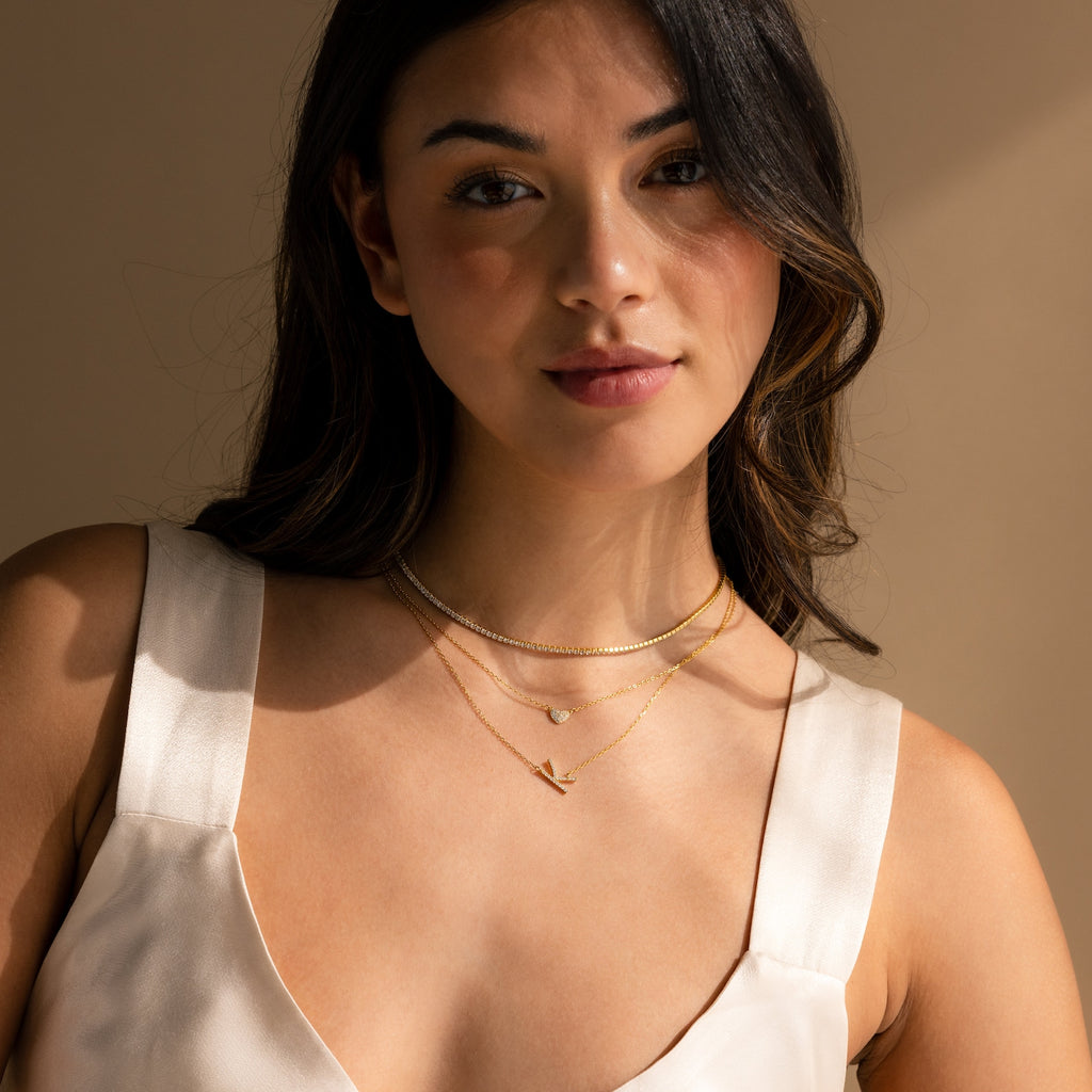 A woman poses against a beige background wearing a cream satin top and the Mini Pave Sideways Initial Necklace in layered gold.