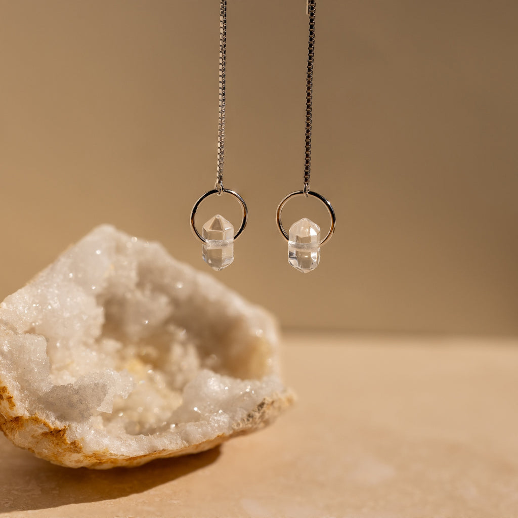 Two crystal pendant necklaces hang beside a large white geode on a beige surface, perfectly complementing Herkimer Diamond Threader Earrings for an elegant, layered look.