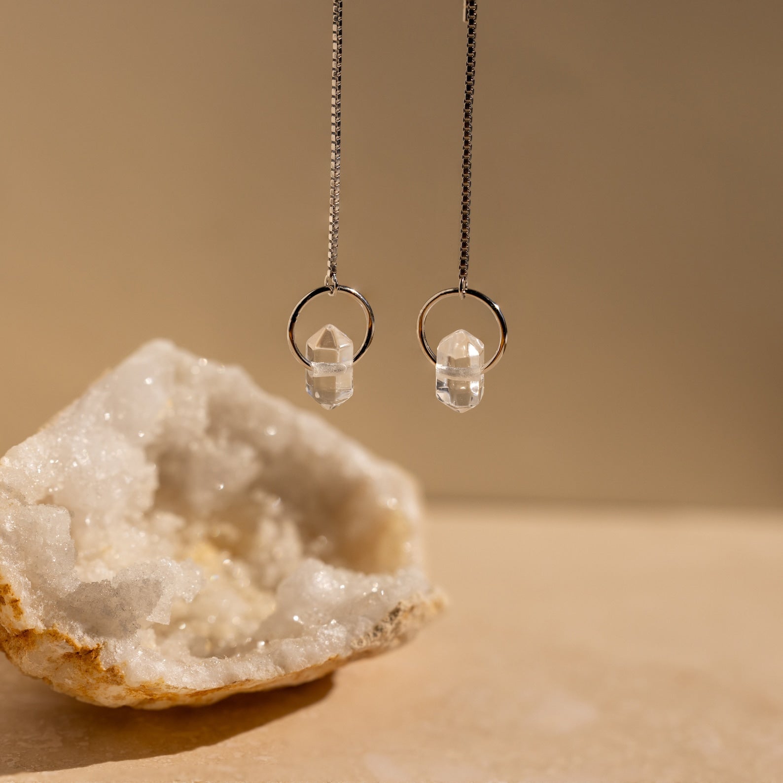 Two crystal pendant necklaces hang beside a large white geode on a beige surface, perfectly complementing Herkimer Diamond Threader Earrings for an elegant, layered look.