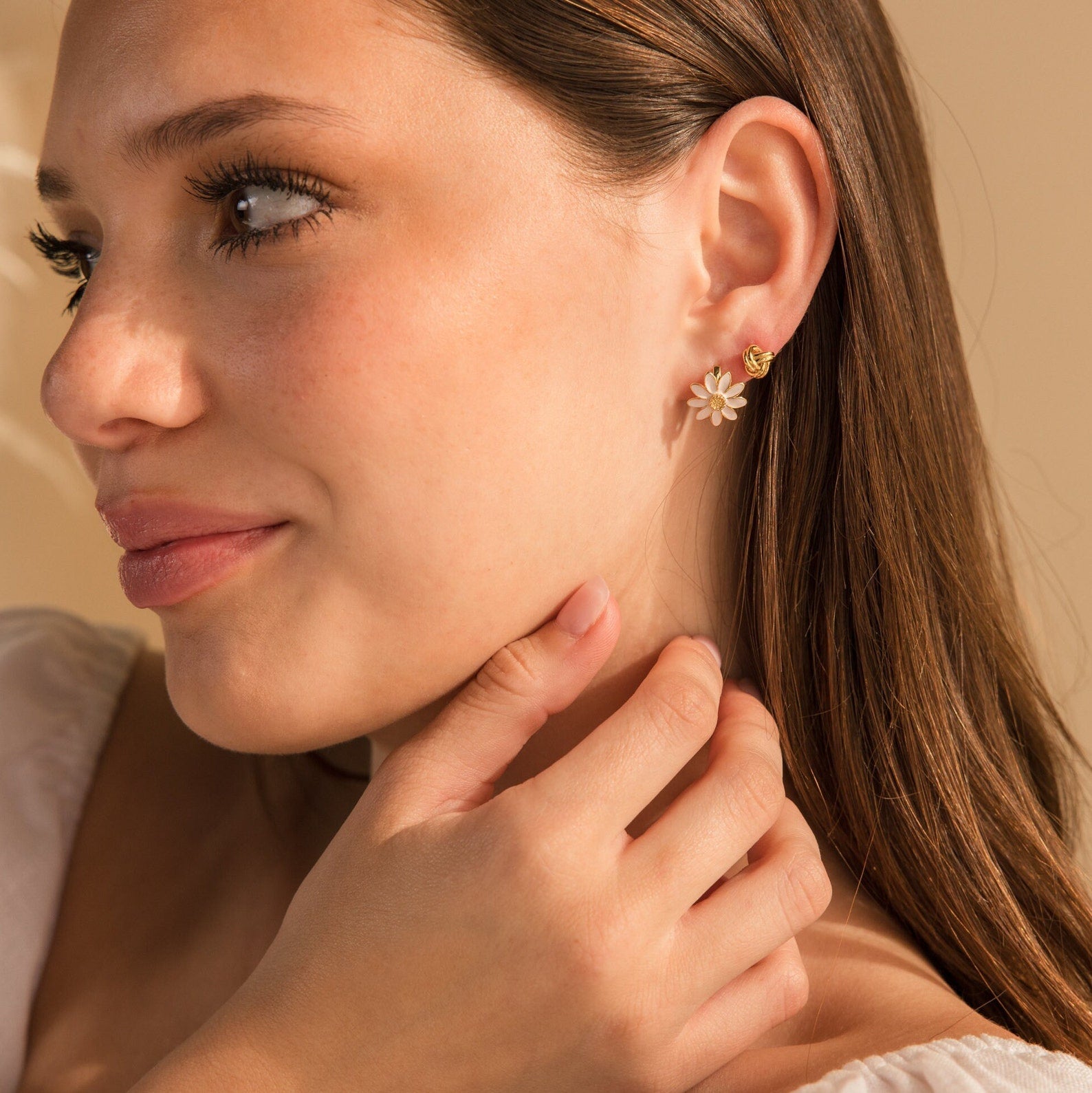 A woman with long brown hair gently touches her neck, elegantly showcasing the minimalist Daisy Huggies while wearing a white top.
