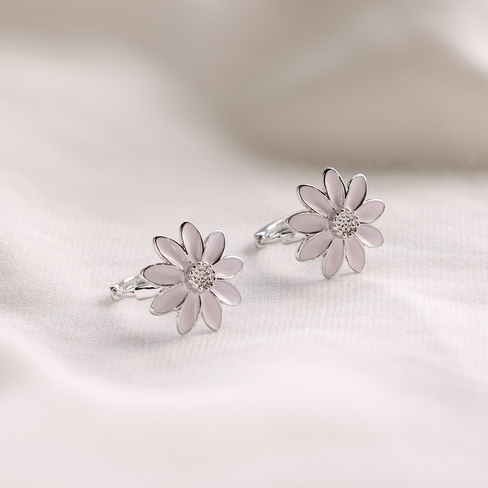 The Daisy Huggies, featuring silver hoops with delicate white petals, are perfect for minimalist jewelry lovers and are shown against a soft, light fabric background.