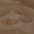 Two sets of gold and silver Mystery Rings from our unique jewelry collection rest on soft beige fabric, with "Mystery Ring" displayed in the center.