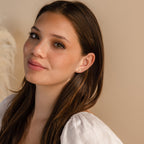 A young woman with long brown hair, wearing a white top and the Marquise Diamond Flower Studs, smiles softly against a beige background.