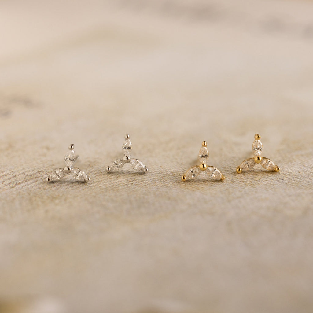 The Trio Floral Marquise Studs set features two pairs of elegant stud earrings in silver and gold, beautifully displayed on a textured beige surface.