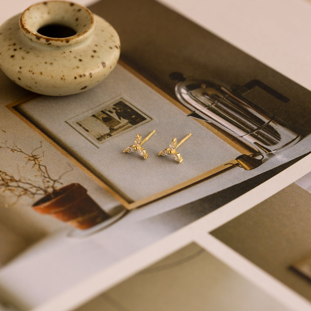 Trio Floral Marquise Studs gold earrings are displayed on an art print, accompanied by a small ceramic vase.