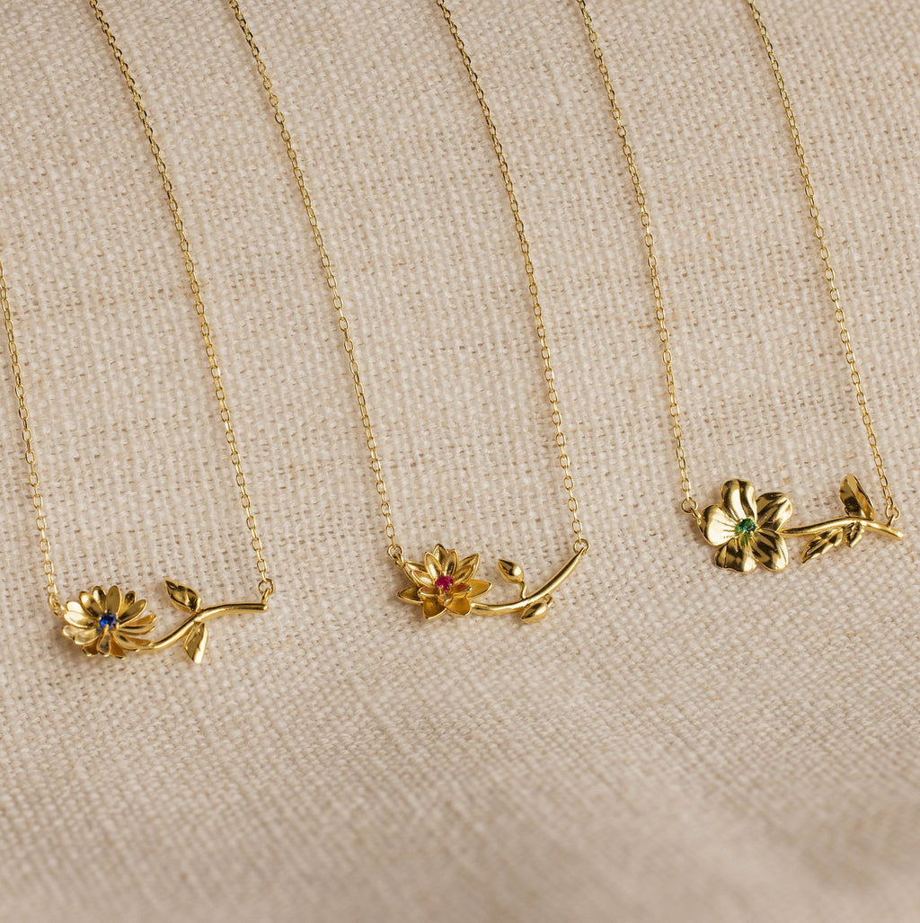 Three Flower Birthstone Necklaces with colored gemstones and personalized charms are displayed on a beige textured fabric background.