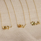 Three Flower Birthstone Necklaces with colored gemstones and personalized charms are displayed on a beige textured fabric background.