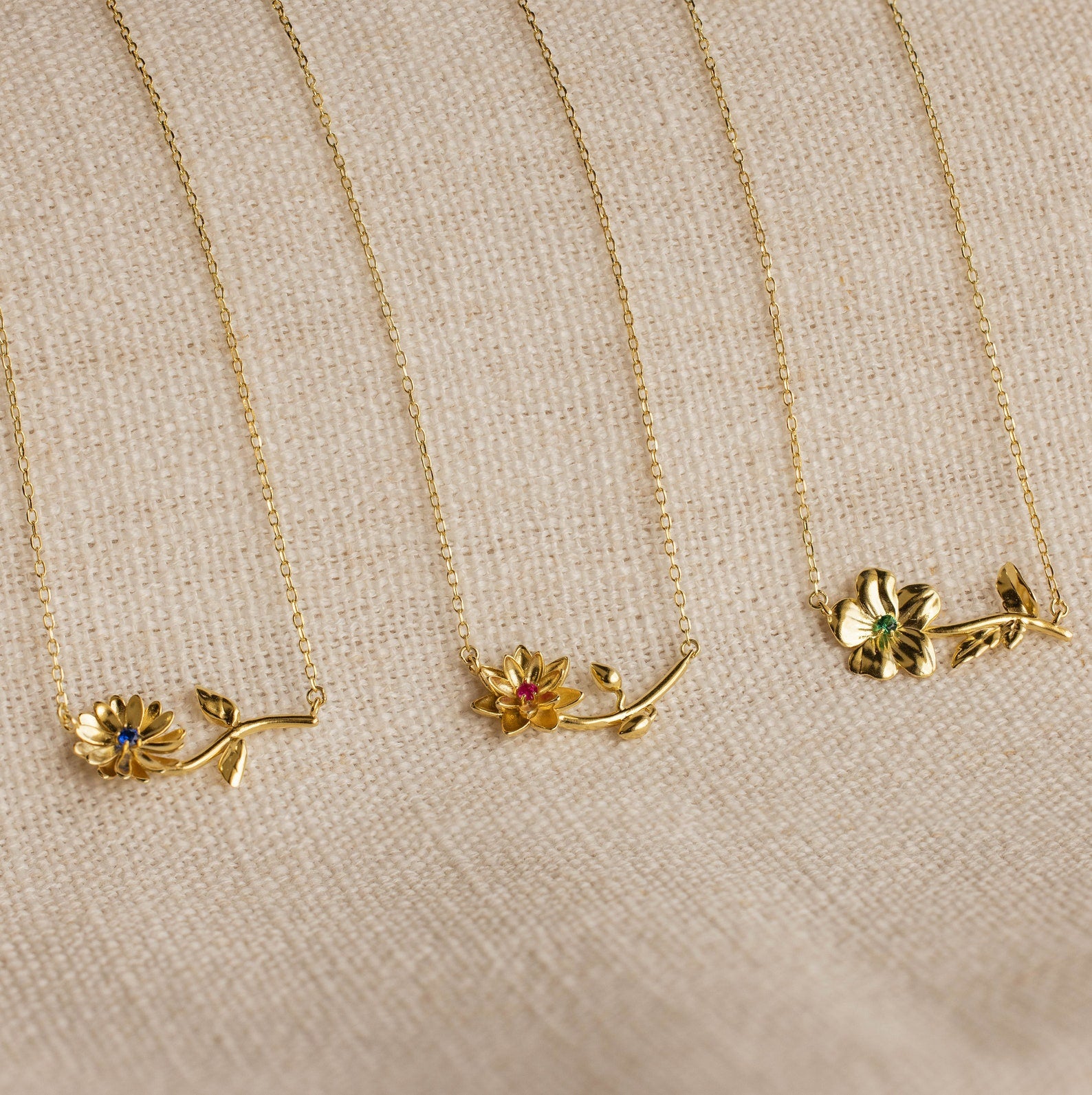 Three Flower Birthstone Necklaces with colored gemstones and personalized charms are displayed on a beige textured fabric background.