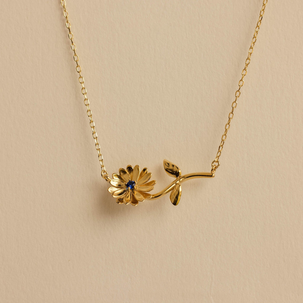 The Flower Birthstone Necklace features a gold chain with a daisy-shaped pendant and a small blue gemstone at its center—ideal for anyone who loves floral jewelry.