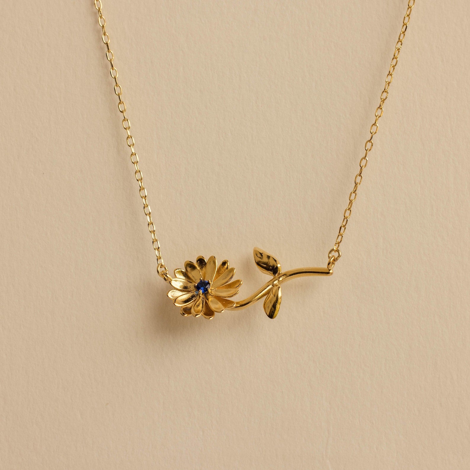 The Flower Birthstone Necklace features a gold chain with a daisy-shaped pendant and a small blue gemstone at its center—ideal for anyone who loves floral jewelry.