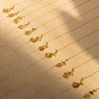 Rows of delicate gold necklaces, such as the Flower Birthstone Necklace, are displayed elegantly on a beige textured surface.