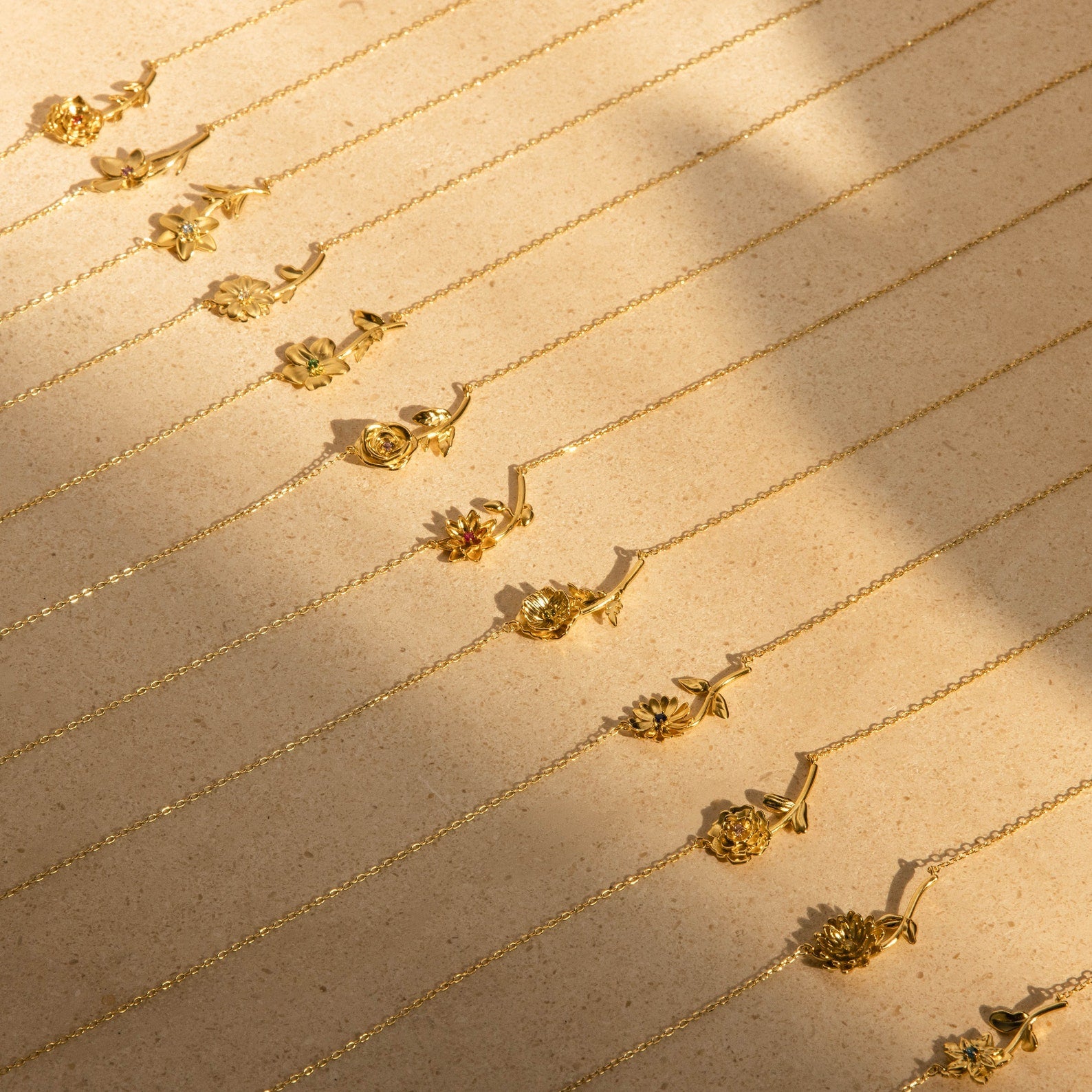 Rows of delicate gold necklaces, such as the Flower Birthstone Necklace, are displayed elegantly on a beige textured surface.