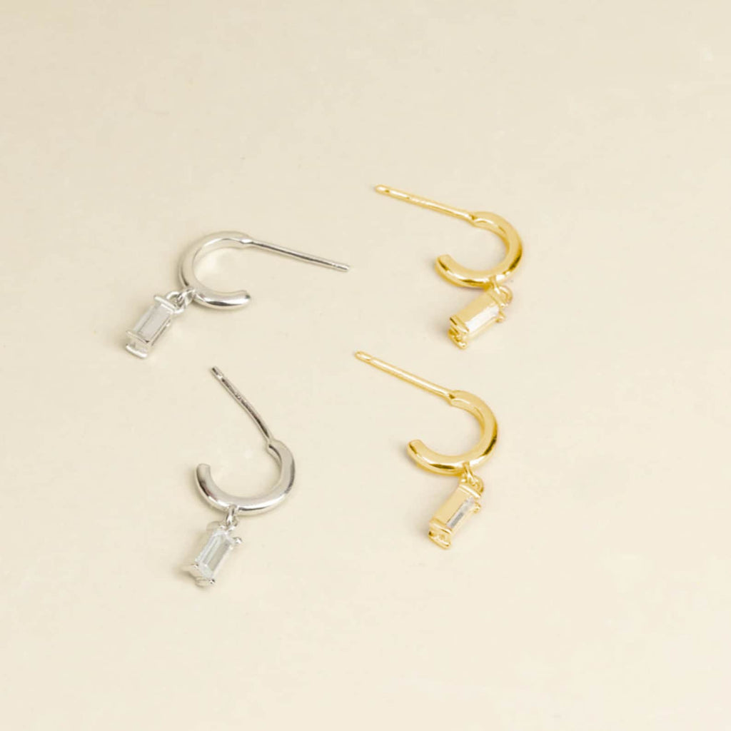 The Lys Baguette Hoops set features two pairs of hoop earrings—one silver, one gold—with rectangular dangling charms, capturing classic elegance against a beige background.