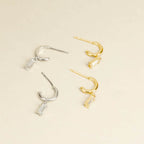 The Lys Baguette Hoops set features two pairs of hoop earrings—one silver, one gold—with rectangular dangling charms, capturing classic elegance against a beige background.