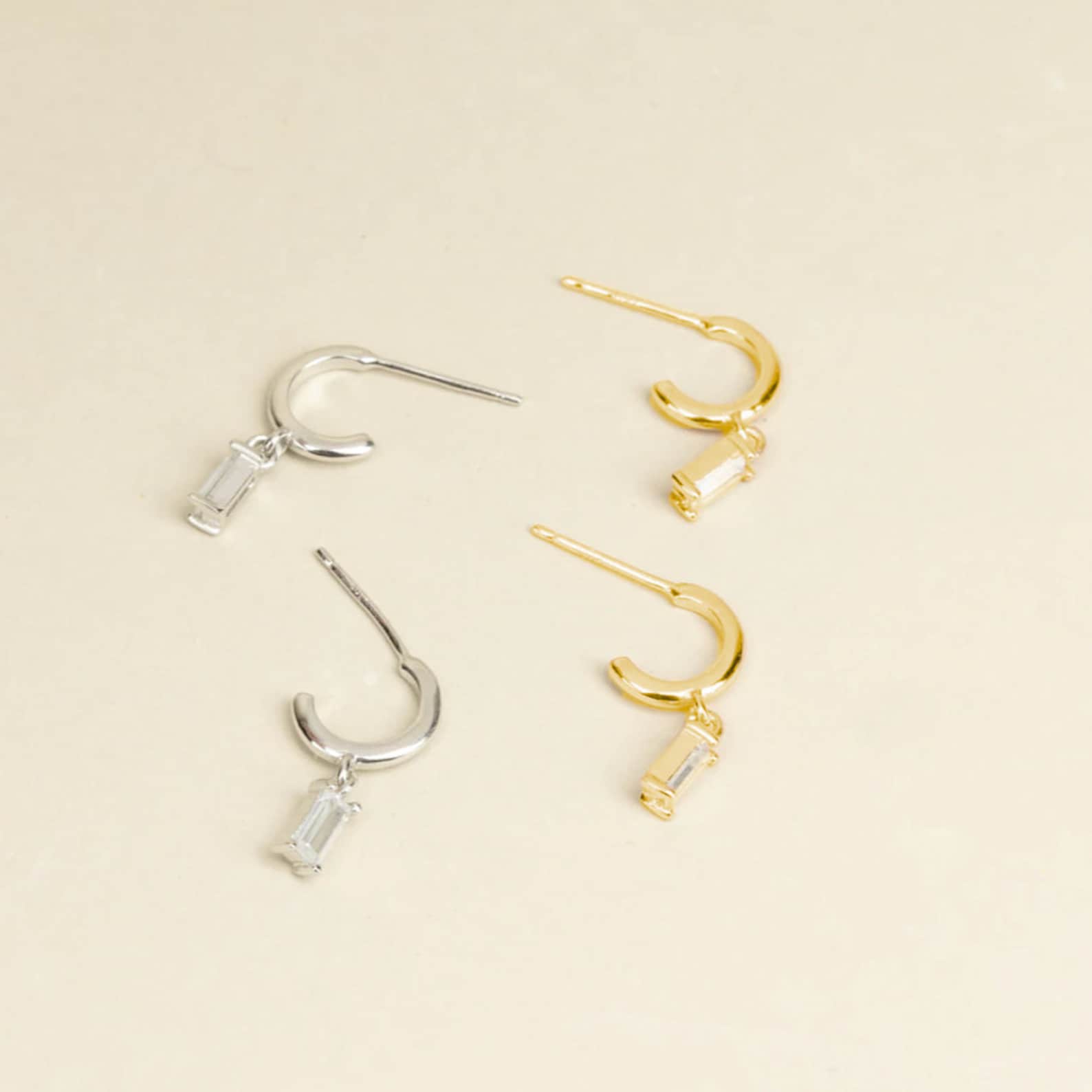 The Lys Baguette Hoops set features two pairs of hoop earrings—one silver, one gold—with rectangular dangling charms, capturing classic elegance against a beige background.