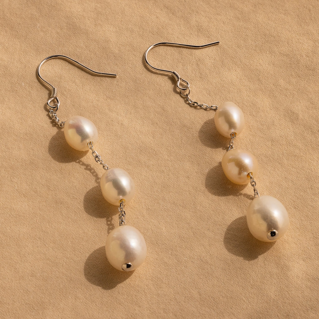 Close-up of a pair of long silver pearl chain earrings set against a tan background.