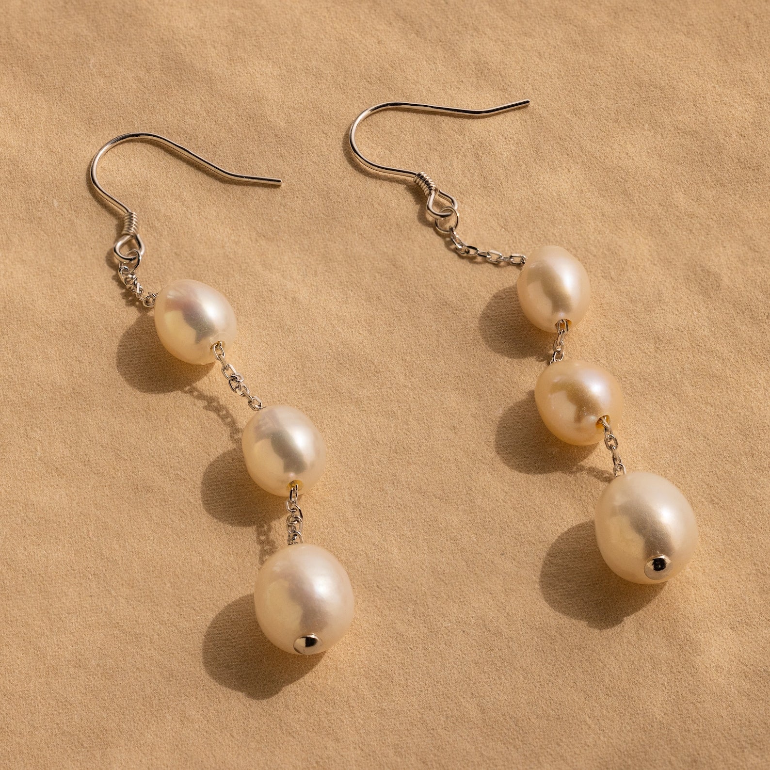 Close-up of a pair of long silver pearl chain earrings set against a tan background.