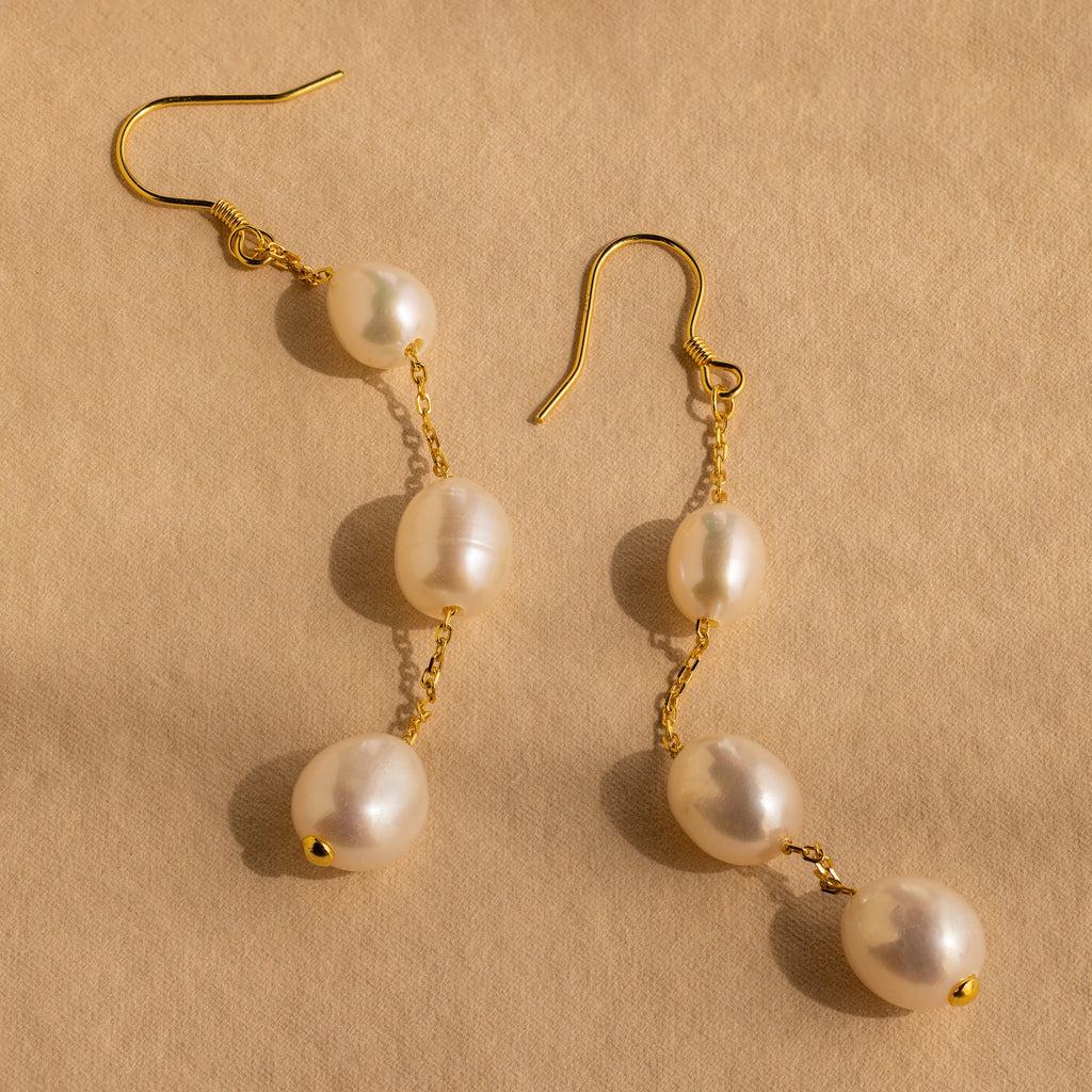 Close-up of a pair of long gold pearl chain earrings set against a tan background.