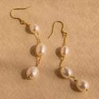 Close-up of a pair of long gold pearl chain earrings set against a tan background.