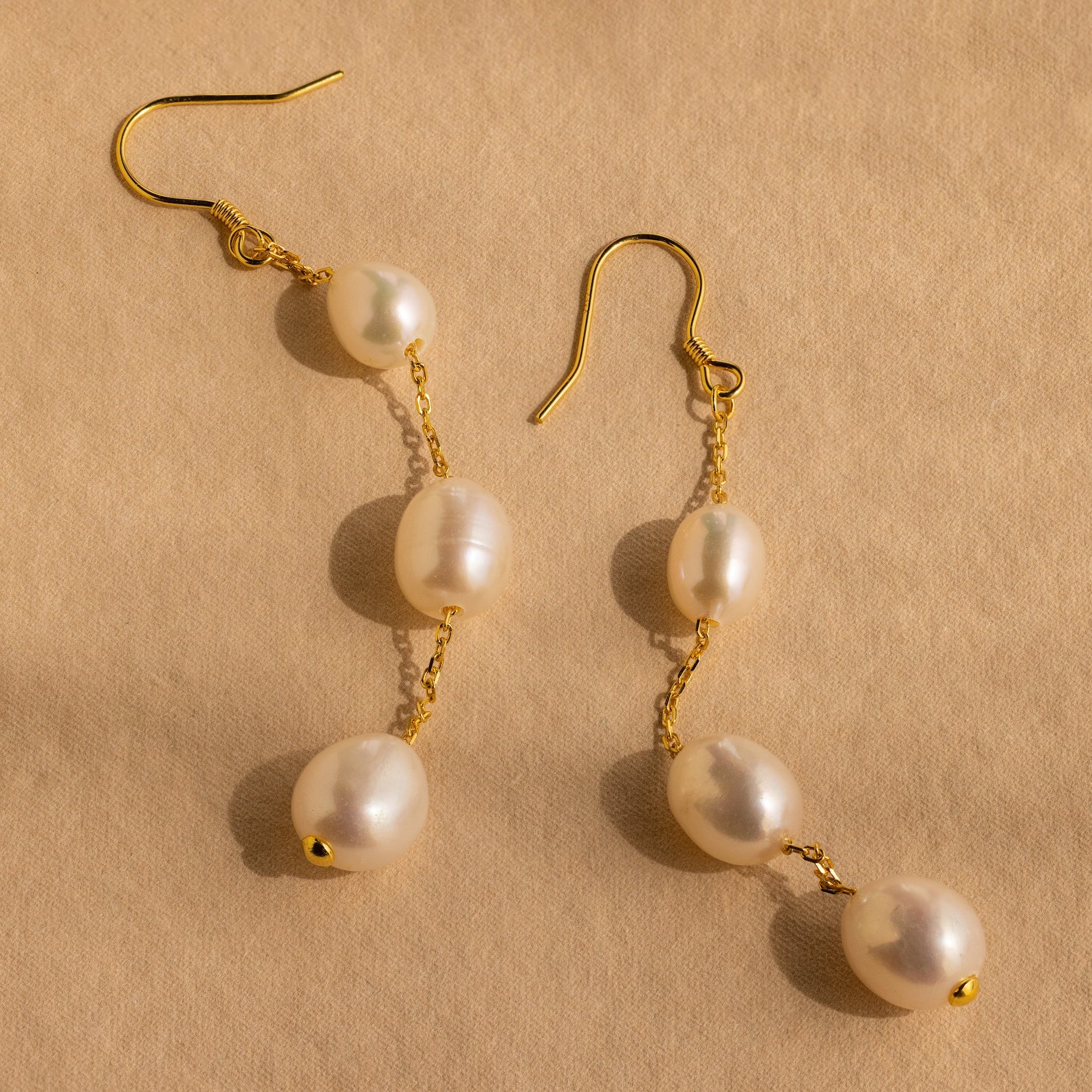 Close-up of a pair of long gold pearl chain earrings set against a tan background.