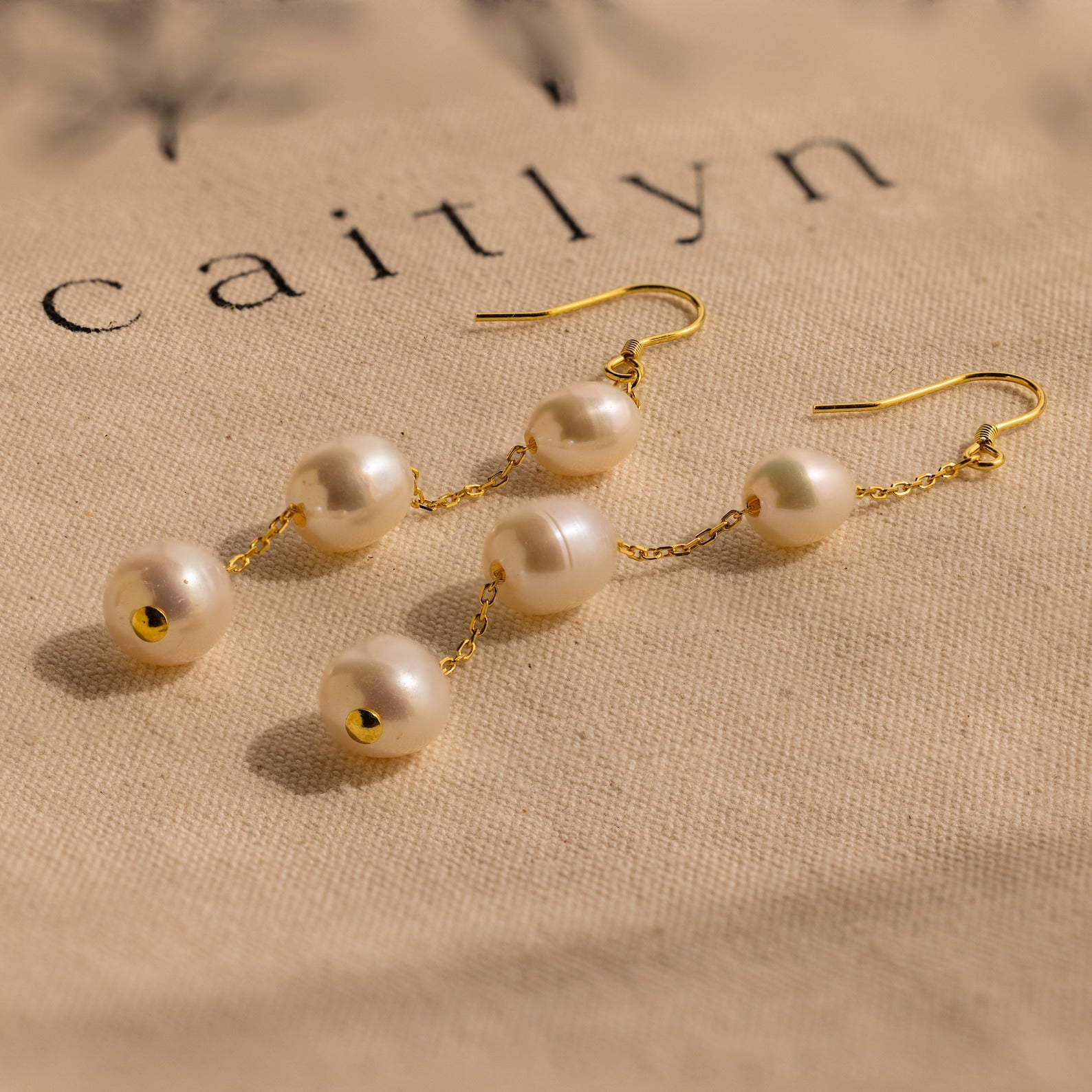 Close-up of long gold pearl chain earrings on beige fabric.