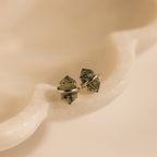 Herkimer Agate Studs: Green crystal stud earrings with silver settings, inspired by geometric gemstone designs, displayed on a smooth, cream-colored surface.