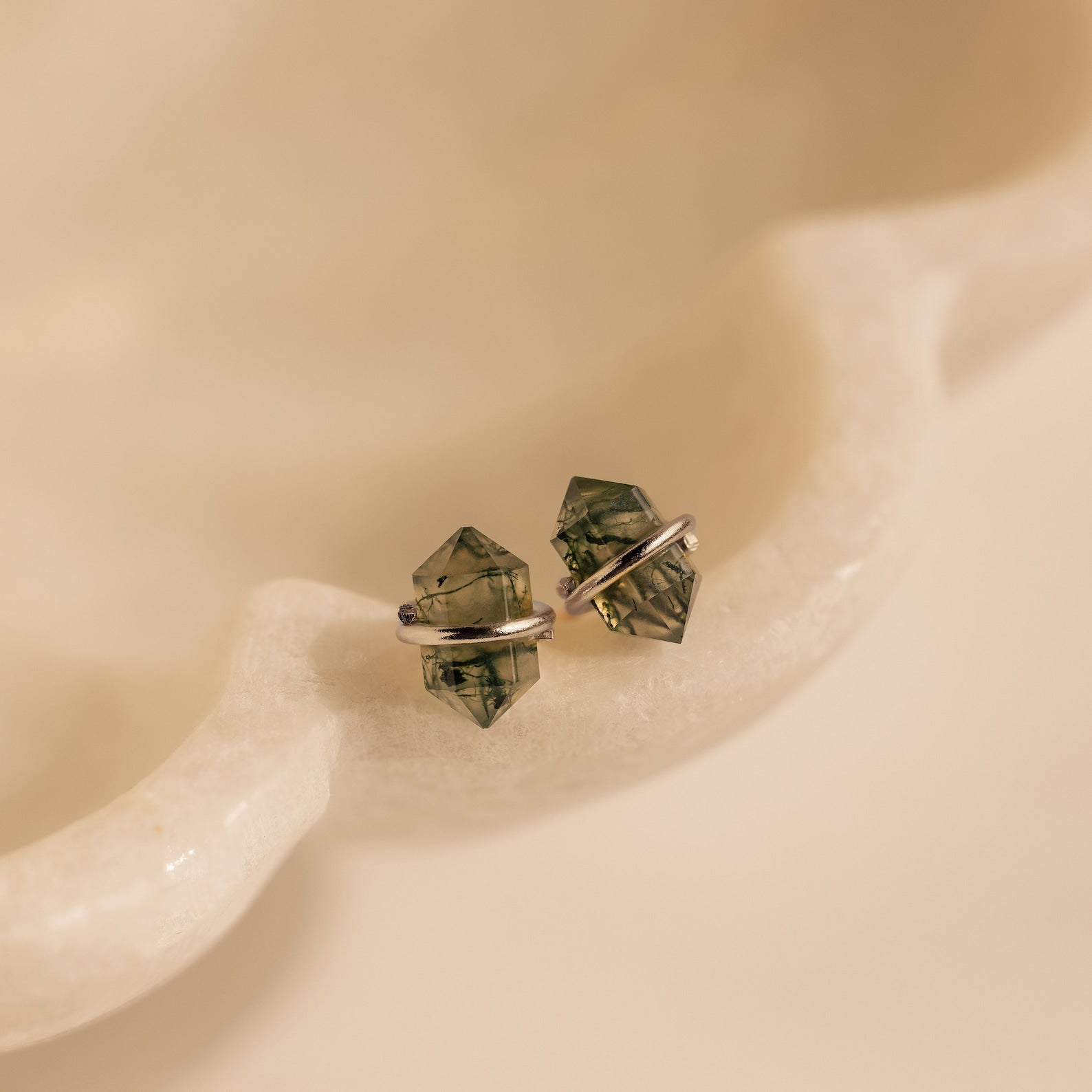 Herkimer Agate Studs: Green crystal stud earrings with silver settings, inspired by geometric gemstone designs, displayed on a smooth, cream-colored surface.