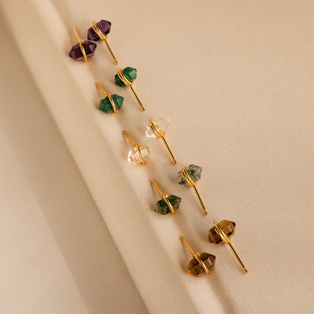 Five pairs of gold geometric gemstone studs, including Herkimer Agate Studs, with a variety of colorful gemstones displayed on beige fabric.