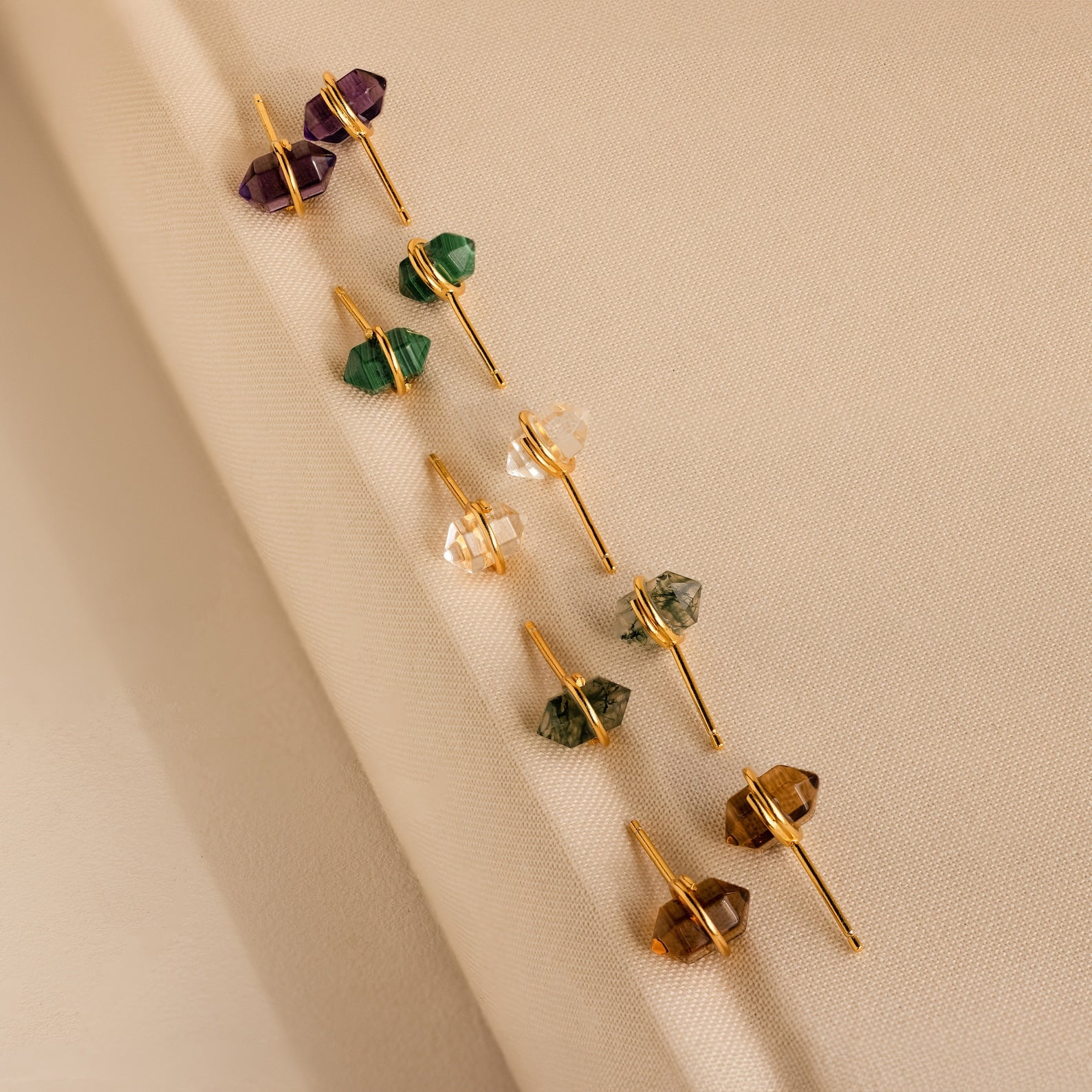 Five pairs of gold geometric gemstone studs, including Herkimer Agate Studs, with a variety of colorful gemstones displayed on beige fabric.