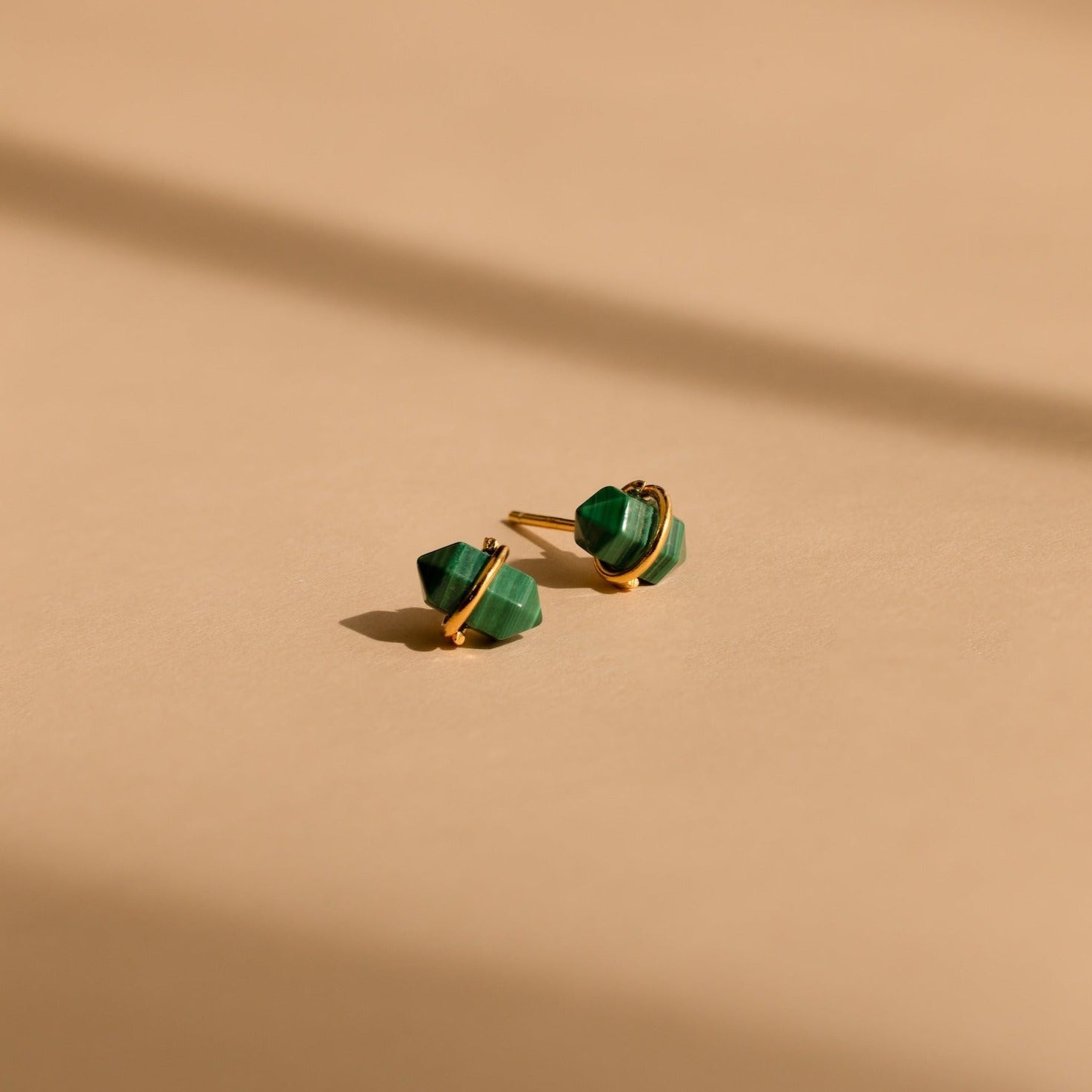 Herkimer Malachite Studs feature gold geometric design with green stone accents, displayed on a beige surface with soft shadows.