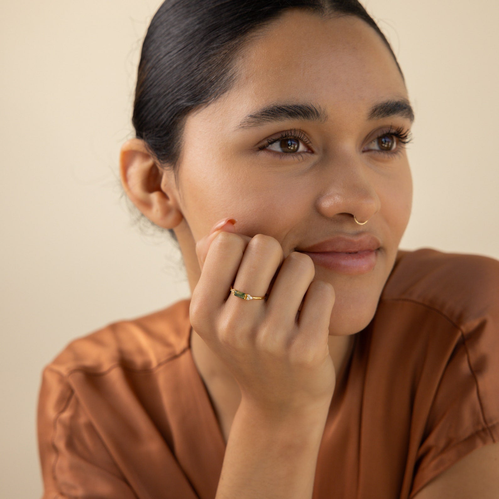 A woman with dark hair and a gold nose ring smiles in a brown top, resting her chin on her hand and subtly showcasing the Agate Baguette Ring as it catches the light.