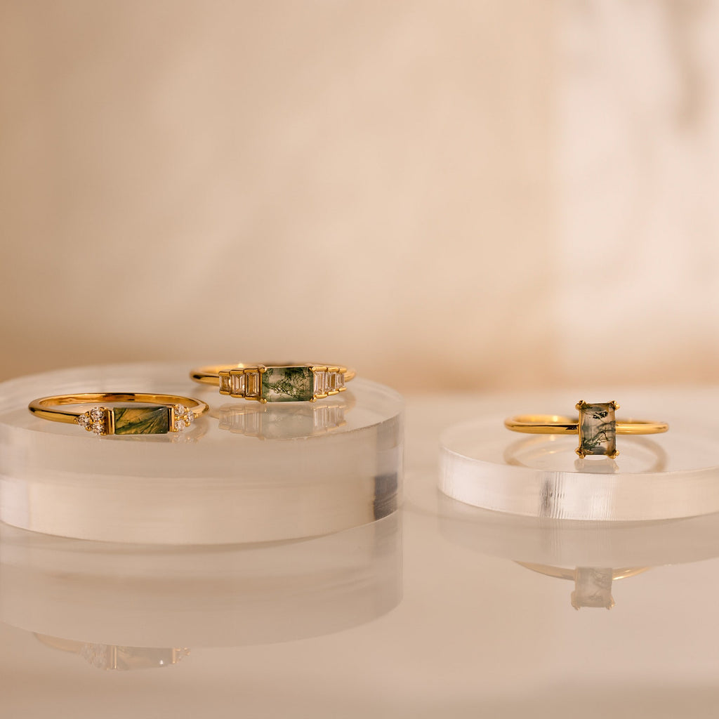 Three gold rings with green gemstone settings, including the Agate Baguette Ring, are displayed on clear, round platforms against a neutral background.