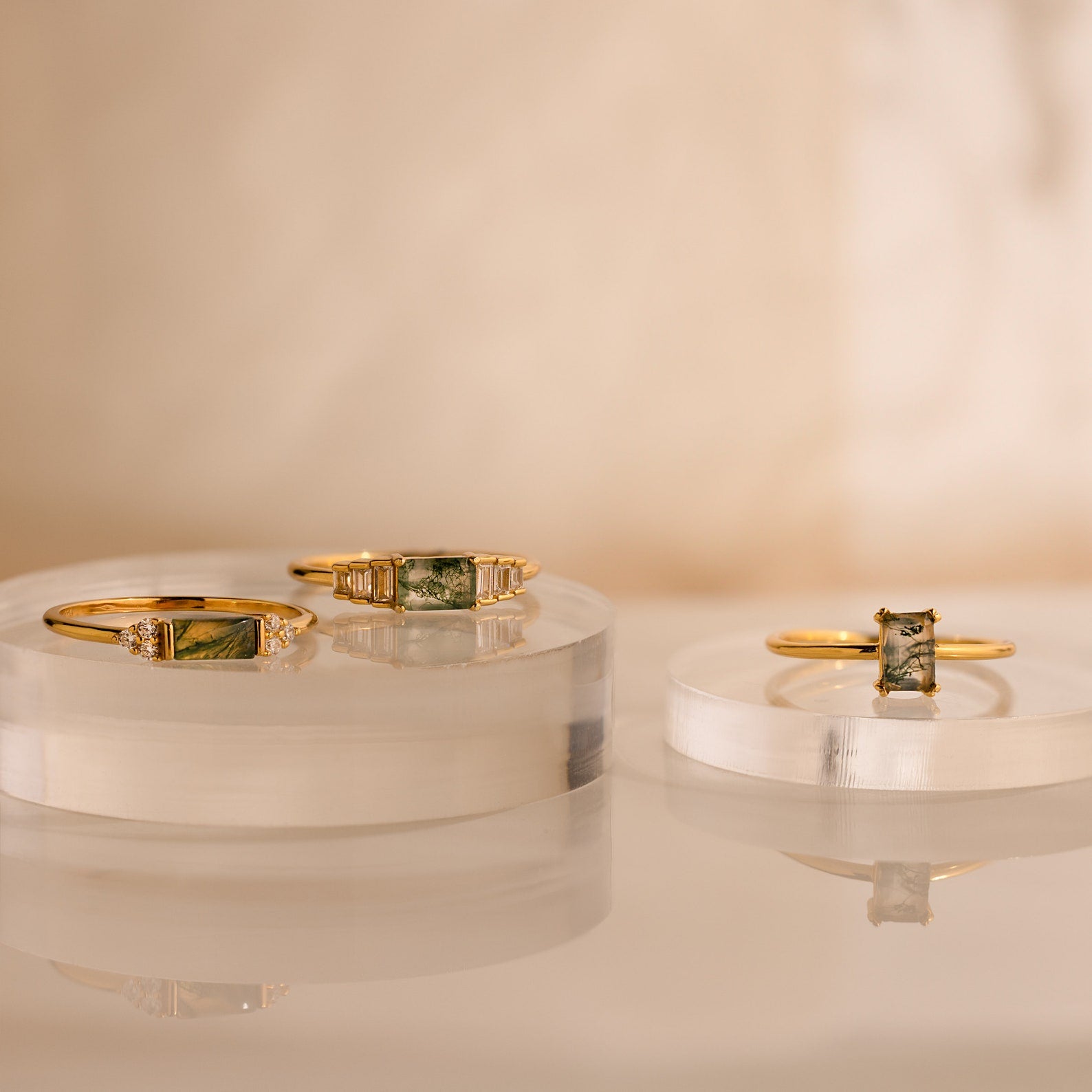 Three gold rings with green gemstone settings, including the Agate Baguette Ring, are displayed on clear, round platforms against a neutral background.