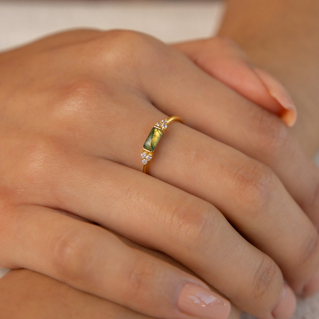 A hand wears the Agate Baguette Ring, featuring a gold band with a green agate stone and small diamond accents, resting gently on another hand.