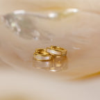 The Mother of Pearl Inlay Huggies rest on a shiny, iridescent surface, highlighting their elegant gold and luminous inlay.