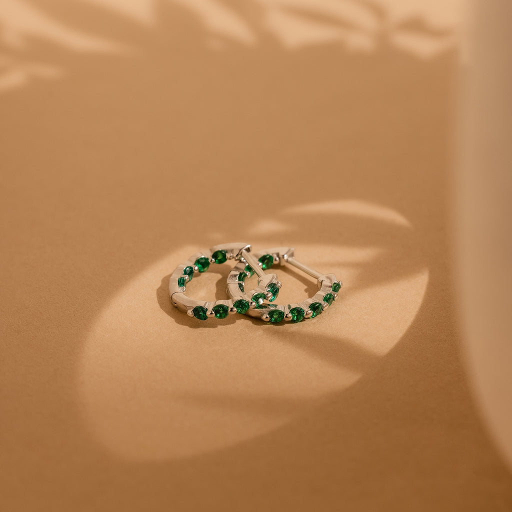 The Inside Out Emerald Huggies, adorned with green stones, exude elegance on a beige surface, beautifully highlighted by soft natural light and gentle shadows.