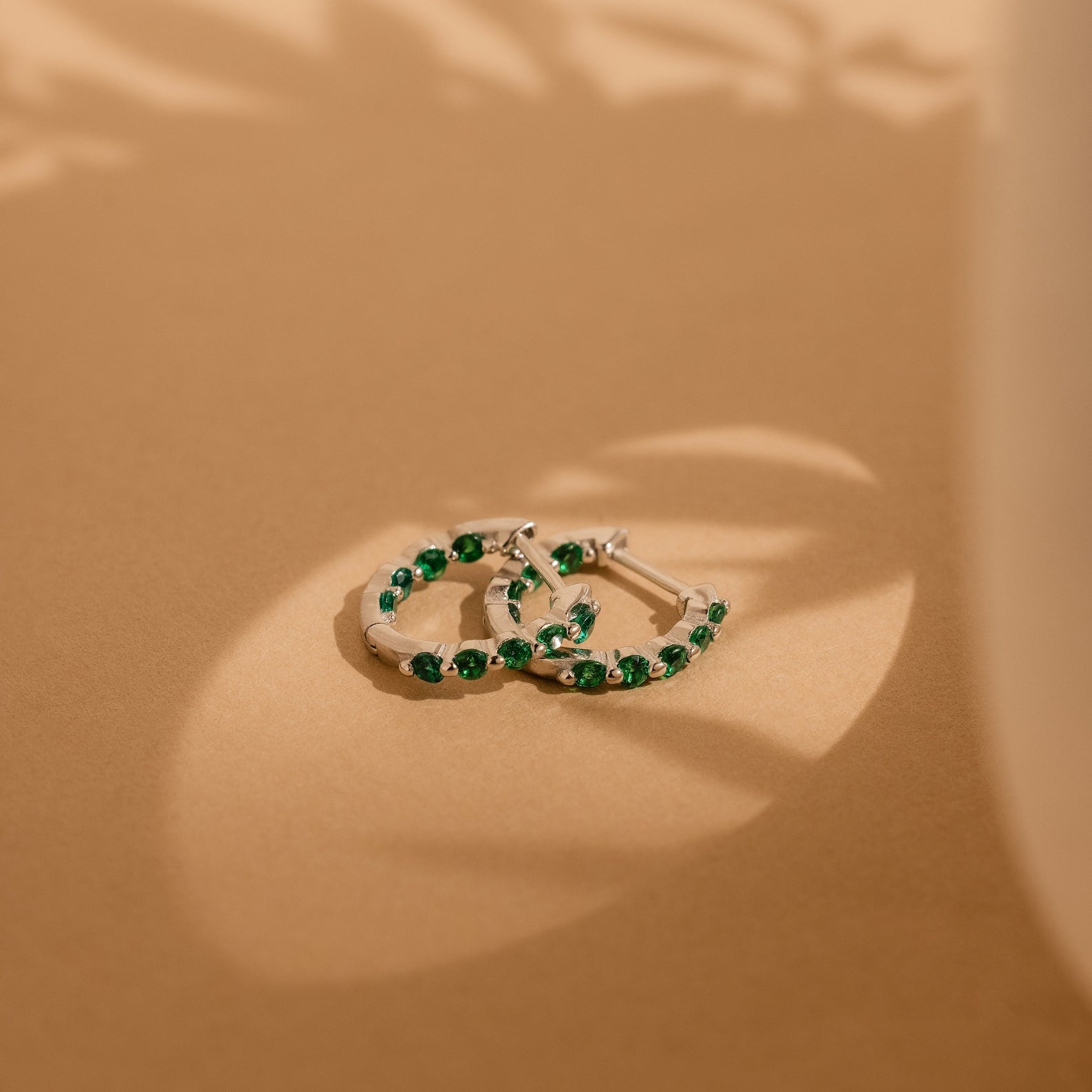 The Inside Out Emerald Huggies, adorned with green stones, exude elegance on a beige surface, beautifully highlighted by soft natural light and gentle shadows.