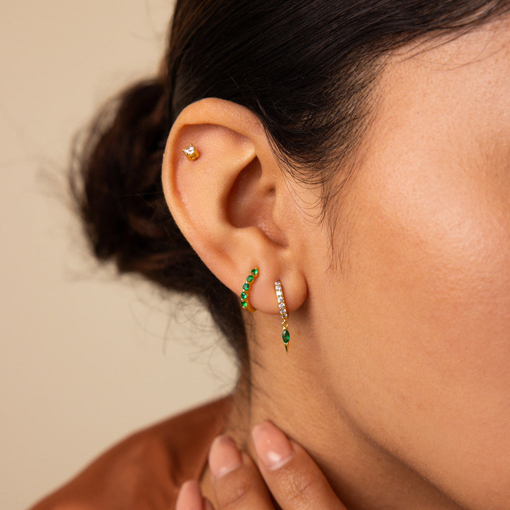 Close-up of a woman's ear with three piercings, featuring the Inside Out Emerald Huggies, a gold earring, and a small stud—an elegant display of modern birthstone jewelry.