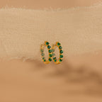 Inside Out Emerald Huggies gold hoop earrings with green gemstones, showcased on textured beige fabric—ideal for birthstone jewelry lovers.