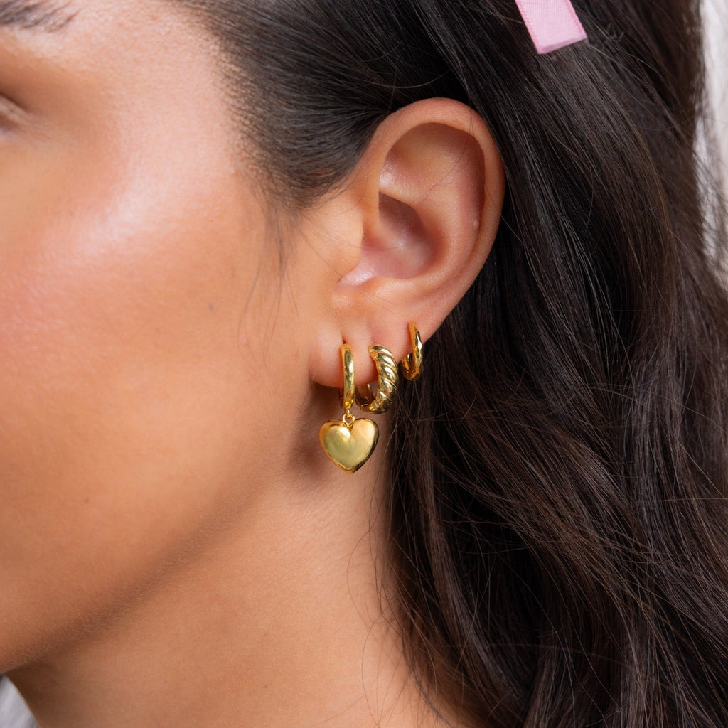 A woman with brown hair and a pink hair clip wears three hoop earrings, including Puff Heart Charm Huggies in Sterling Silver.
