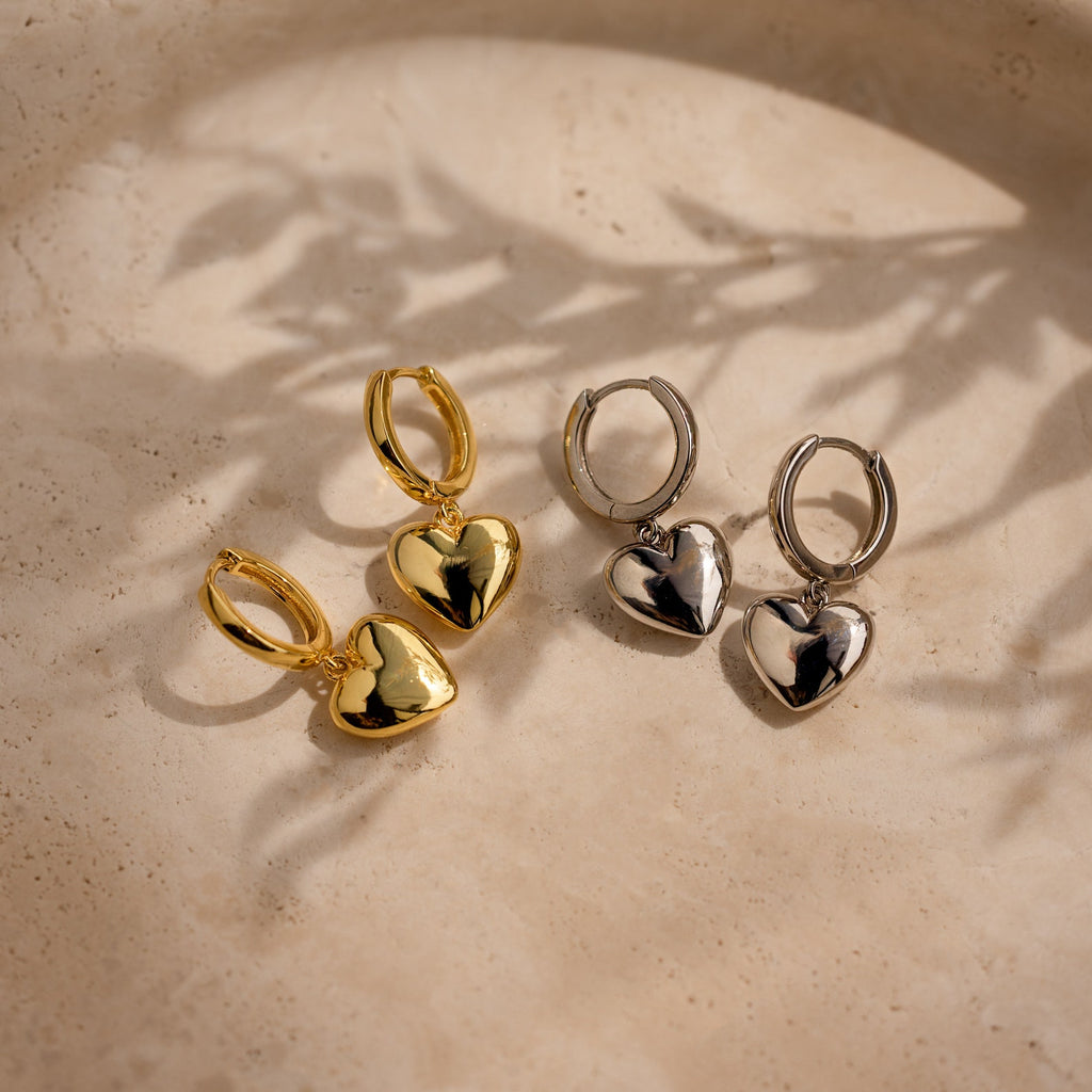A pair of Puff Heart Charm Huggies in 18K Gold with a dainty heart-shaped design, displayed on a beige textured surface.
